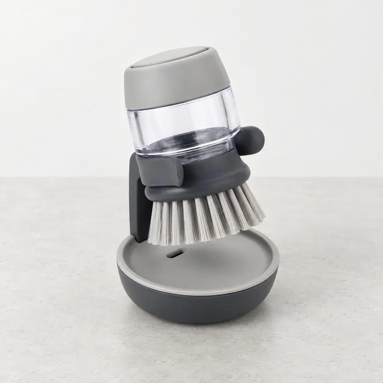 Grey soap-dispensing palm brush with a clear soap reservoir standing on its charcoal drip-dry stand.