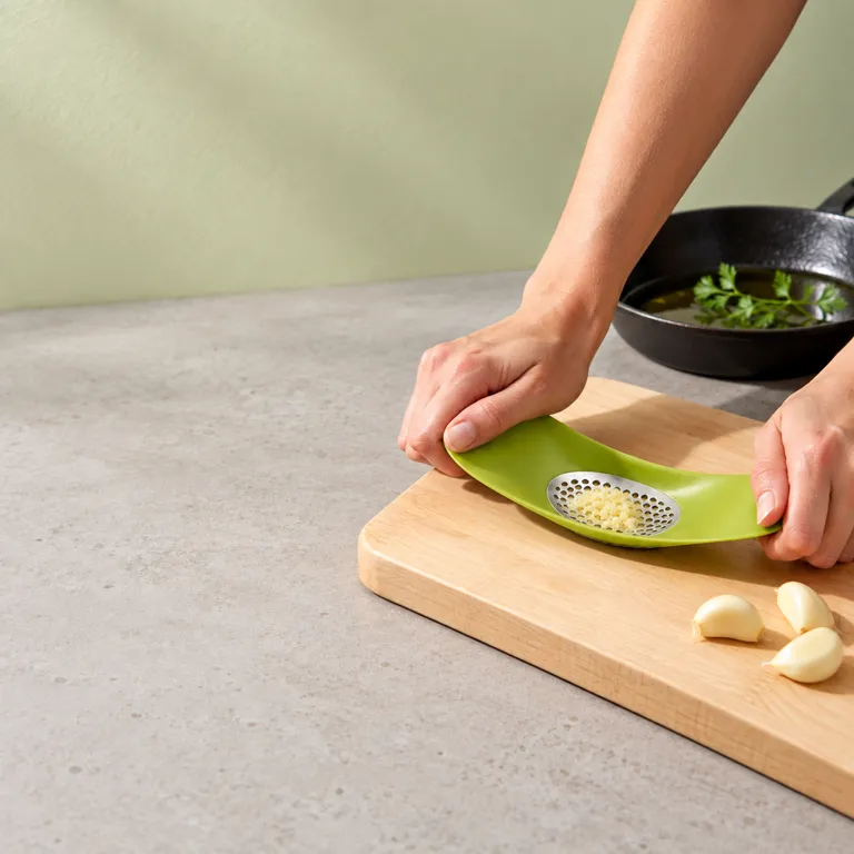 Two hands pressing the green garlic crusher over peeled cloves on a birch board, with a skillet waiting behind.