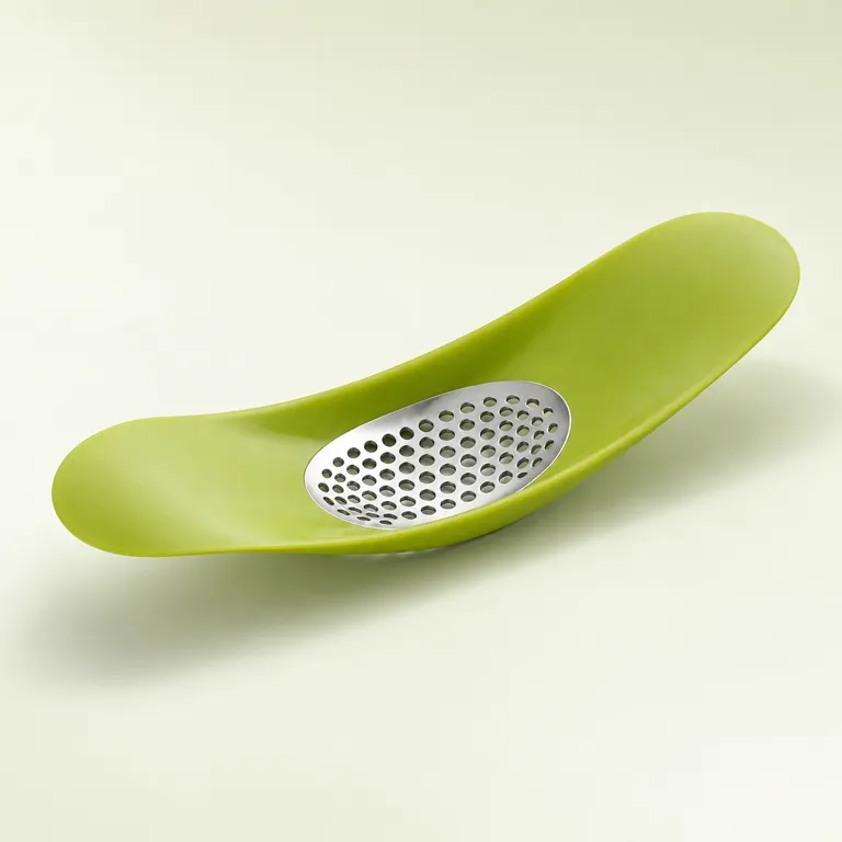 A lime-green curved garlic crusher with an oval perforated plate in its bowl-shaped top, on a pale sage surface.
